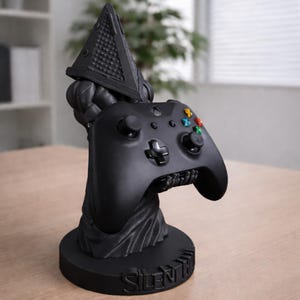 May include: A black Xbox controller is held by a black Pyramid Head-themed stand. The stand features a pyramid-shaped head and the words "Silent Hill" on the base. The controller has coloured buttons and a directional pad.