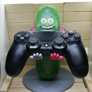 May include: A green pickle-shaped video game controller holder with a smiling face, holding a black PlayStation controller. The holder has pink and gray hand-like supports and sits on a gray circular base.