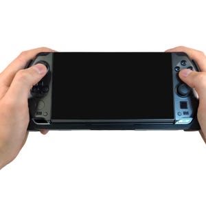 May include: A handheld gaming console with a black screen and gray controllers. The console is held by two hands, with thumbs on the control sticks and buttons. The device has a sleek, modern design.