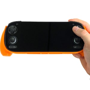 May include: A handheld gaming device with an orange grip and black screen. The device features a directional pad, buttons, and analog sticks. The screen is black, and the device is held by a person's hand.