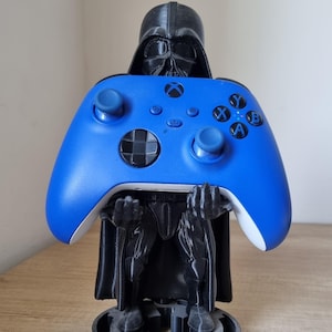 May include: A blue Xbox controller is held by a black Darth Vader-themed stand. The controller has blue buttons and directional pads. The stand is 3D-printed and features the iconic helmet and armour of the Star Wars character.