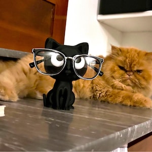 Puede incluir: Un soporte para gafas con forma de gato negro con gafas con montura plateada. El gato está sentado sobre una superficie gris. Un gato naranja está en el fondo.