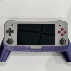 May include: A handheld gaming console in a gray case with a black screen, a directional pad, and pink buttons. The console is held in a purple grip with the brand name "ANBERNIC" visible.