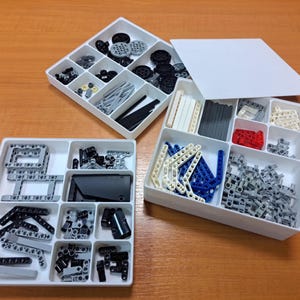 May include: Three white plastic storage containers filled with various small, black, grey, white, and blue building toy pieces. The pieces include gears, rods, connectors, and other components, organised in separate compartments.
