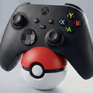 May include: A black Xbox controller sits atop a red, white, and black Pokeball. The controller features coloured buttons and two joysticks. The Pokeball is round with a white base and a red top.