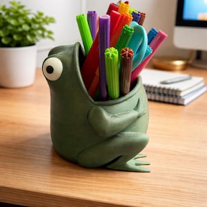 May include: A green frog-shaped desk tidy filled with colourful markers and pens. The frog has a large, expressive eye and is sitting on a wooden desk. The organiser is a fun and functional way to store writing utensils.