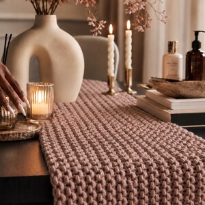 May include: A dining table setting with a mauve knitted table runner. The table is decorated with a vase of flowers, candles, and decorative bottles. A hand is reaching towards a perfume bottle on a wooden coaster.