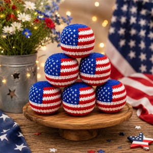 May include: Six crocheted balls, each with a design inspired by the American flag. The balls are blue with white stars and red and white stripes. They are arranged on a wooden cake stand, with a patriotic theme. The balls are approximately 7 cm in diameter.