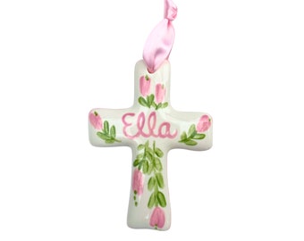 May include: White ceramic cross with pink flowers and green leaves. The name "Ella" is painted in pink on the cross.