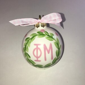 May include: White ceramic Christmas ornament with a pink and green wreath design. The wreath encircles the letters "ΦΜ" in pink.