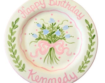 May include: A white ceramic plate with a pink border and a floral design. The plate has the words "Happy Birthday" and "Kennedy" written in pink.