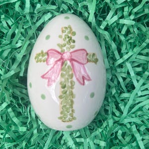 May include: White Easter egg decorated with a cross made of green leaves and a pink bow. The egg has green polka dots and is set on a bed of green shredded paper. A festive, handmade Easter decoration.