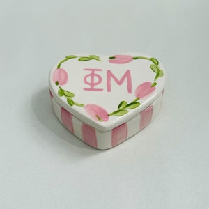 May include: Heart-shaped ceramic trinket box with a white base and pink stripes. The lid features pink tulips and green leaves around the edge, with the Greek letters Φ and M in pink in the center.
