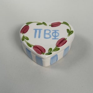 May include: Heart-shaped ceramic trinket box with a white lid featuring a wreath of red tulips and green leaves. The box has blue vertical stripes on the sides and Greek letters in blue. A decorative storage container.