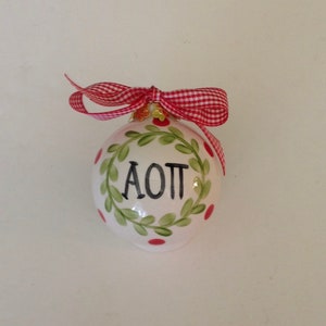 May include: A white ceramic Christmas ornament with the letters "AOTT" in black, encircled by a green wreath with red berries. A red and white checkered ribbon is tied at the top.