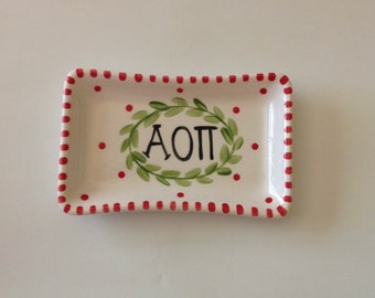 May include: A white ceramic dish with a red and white striped border and red polka dots. The dish has a green wreath with the letters "AOP" in the centre.