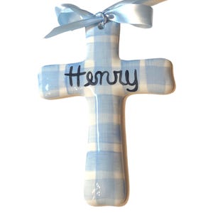 May include: A ceramic cross ornament with a blue and white gingham pattern. The word "Henry" is written in black script across the center. A light blue satin ribbon is tied at the top for hanging.