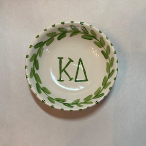 May include: A white ceramic bowl with a green leaf design around the rim and the letters "KΔ" in the center. The bowl is round and appears to be hand-painted, with a simple and elegant aesthetic.