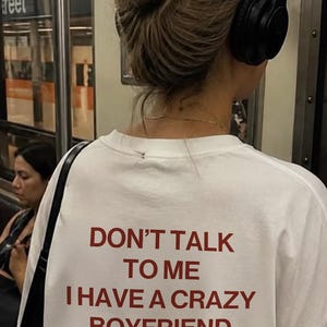 Peut inclure: Un t-shirt blanc avec le texte rouge DON'T TALK TO ME I HAVE A CRAZY BOYFRIEND imprimé au dos. Sous le texte, le mot Remarkz est écrit dans une police rouge cursive plus petite. Le t-shirt a un col rond et des manches courtes.