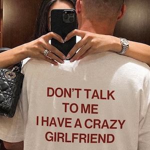 Könnte beinhalten: Ein weißes kurzärmeliges T-Shirt mit dem dunkelroten Aufdruck DON'T TALK TO ME I HAVE A CRAZY GIRLFRIEND auf der Rückseite, wobei das Wort Remarkz in einer kleineren Schreibschrift unter dem Haupttext gedruckt ist.