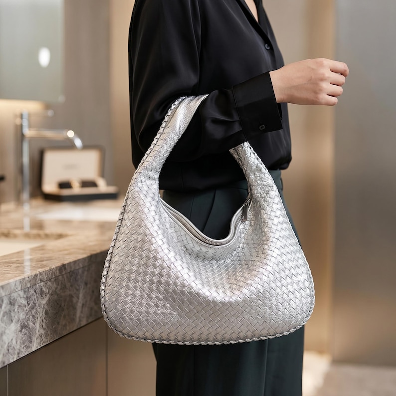 May include: A silver woven leather hobo bag with a curved shape and a zipper closure. The bag has a wide, comfortable strap and is held by a person wearing a black shirt and dark pants. The bag's metallic sheen catches the light.