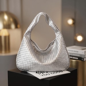 May include: Silver woven shoulder bag with a curved handle. The bag has a textured appearance and a smooth, shiny finish. The bag is sitting on a black surface, with a white card that says "YOU GOOD".