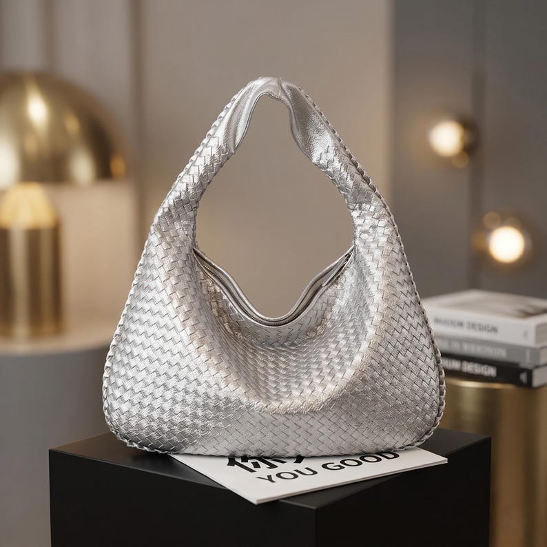 May include: Silver woven shoulder bag with a curved handle. The bag has a textured appearance and a smooth, shiny finish. The bag is sitting on a black surface, with a white card that says "YOU GOOD".
