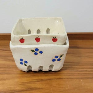 May include: Two stacked, off-white ceramic berry baskets. The top basket has red apple designs, while the bottom basket features blue blueberry designs. Both have a rustic, handmade appearance and are set on a wooden surface.