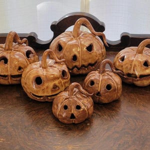 May include: A collection of six brown glazed ceramic jack-o'-lanterns of varying sizes. Each pumpkin features a carved face with cut-out eyes, noses, and mouths, topped with a curved stem. The glossy finish reflects light across the surfaces.