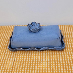 May include: A blue ceramic butter dish with a flower-shaped handle on the lid. The dish has a rectangular shape with a wavy edge and sits on a yellow and white checkered tablecloth.
