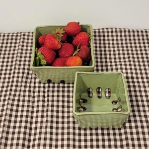 May include: Two square, light green ceramic berry baskets with perforated sides and bottoms. One basket is filled with fresh, red strawberries, while the other empty basket sits in front, showcasing the glossy finish and cutout design.