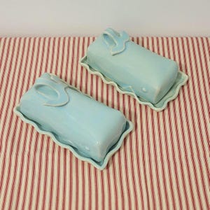 May include: Two rectangular light blue ceramic butter dishes with matching lids. Each lid features a sculpted handle on top, and both the base and lid have a wavy, scalloped edge design. The ceramic has a smooth, glossy finish throughout.