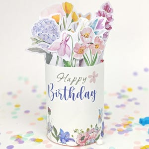 May include: A cylindrical pop-up greeting card featuring a bouquet of watercolor-style flowers in shades of blue, pink, yellow, and purple. The white exterior displays the text Happy Birthday in blue script with a small pink flower icon above it.