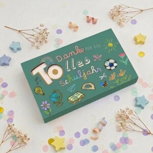 Pode incluir: Uma caixa de presente retangular verde com o texto "Tolles Danke für ein Schuljahr" em letras coloridas. A caixa é decorada com ilustrações de um globo, um livro, um transferidor, uma abelha, um sol, flores e um arco-íris.