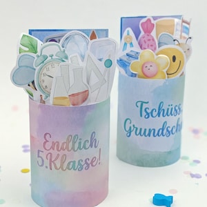 Può includere: Due contenitori cilindrici in carta con sfondi pastello in stile acquerello. Uno riporta il testo Endlich 5.Klasse! e l'altro Tschüss Grundschule. Entrambi sono riempiti con vari ritagli di carta a tema scolastico, tra cui un orologio, becher, un fiore e una faccina sorridente.