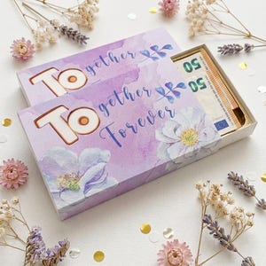 Könnte beinhalten: Eine rechteckige Geschenkbox mit violettem Aquarell-Blumenmuster und dem Text "Together Forever" in weißer und blauer Schrift. Die Box ist leicht geöffnet und zeigt einen Stapel 50-Euro-Scheine im Inneren, umgeben von getrockneten Blumen.