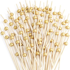 Op de afbeelding: Een verzameling bamboe cocktailprikkers, elk voorzien van een gladde, metallic goudkleurige bolvormige kraal nabij de bovenkant. De prikkers zijn in een waaier gerangschikt, waarbij de scherpe uiteinden en de lichtgekleurde houten schachten zichtbaar zijn.