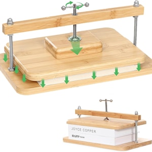 May include: A bamboo book press featuring two rectangular wooden boards, four threaded metal rods with nuts, and a central screw mechanism with a handle. The device is shown compressing two white books labeled JOYCE COPPER and HAPPINESS.