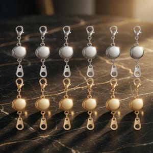 May include: Six zipper pull charms with a silver and gold color scheme. Each charm features a round, metallic bead, a zipper-shaped charm, and a lobster clasp. The charms are displayed on a dark, marble-like surface.