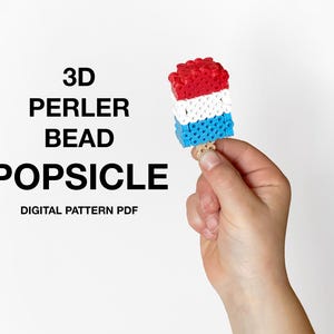 May include: A 3D popsicle constructed from red, white, and blue Perler beads with a small wooden stick at the base. The text 3D PERLER BEAD POPSICLE DIGITAL PATTERN PDF is printed in black on the left side of the image.