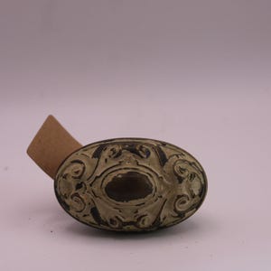 May include: An oval-shaped cabinet knob featuring a distressed cream-colored finish with dark brown recessed detailing and intricate scrollwork patterns. A small, blank brown cardboard tag is attached to the side of the metal hardware piece.