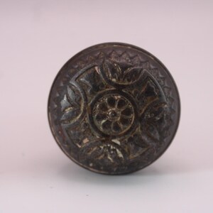 May include: A circular metal door knob with a dark, weathered finish. The surface features a central floral medallion surrounded by four symmetrical leaf-like motifs, all enclosed within a decorative border with a repeating serrated edge pattern.