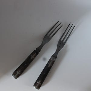 May include: Two metal forks with dark, rectangular handles. Each handle features two inlaid cross symbols in a lighter metallic tone. The forks have four tines each and a metallic finish that appears slightly weathered or aged in appearance.