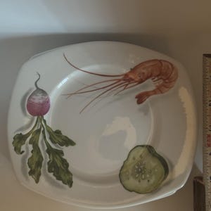 May include: A white square ceramic plate with rounded corners featuring a painted illustration of a pink shrimp, a purple and white radish with green leaves, and a sliced green cucumber. A wooden ruler is visible on the right side for scale.