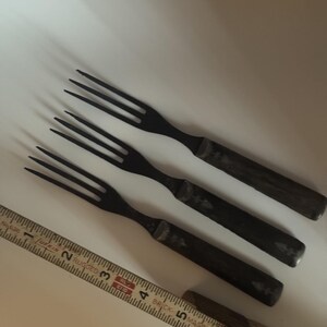 May include: Three black forks with four tines and rectangular handles, each featuring a small etched tree design. A yellow measuring tape is positioned alongside the forks, showing a length of approximately 6.5 inches for the cutlery items.