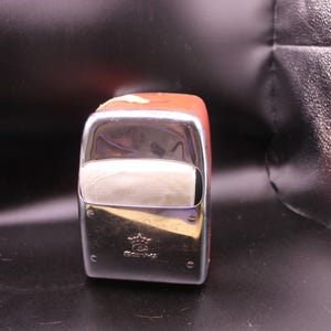 May include: A rectangular napkin dispenser with a polished chrome front panel featuring a small crown logo and the text "GUEST" embossed at the bottom. The dispenser has a reddish-brown body and a central opening for dispensing paper napkins.