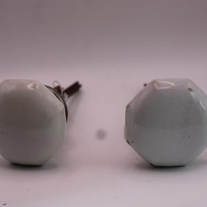 May include: Two white octagonal ceramic door knobs with a smooth, glossy finish. Each knob features a central metal spindle extending from the back, designed for attachment to a door mechanism. The surface of the ceramic shows minor surface wear.