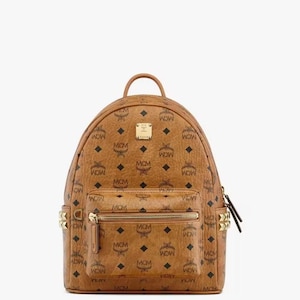 Op de afbeelding: Een bruine rugzak met een herhalend MCM-logopatroon. De rugzak heeft een handvat aan de bovenkant, een voorvak met ritssluiting en goudkleurige stud-accenten. De tas heeft een goudkleurige rits en een kleine goudkleurige logoplaat.