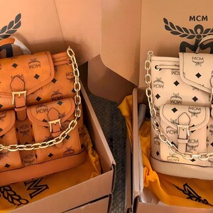 以下が含まれることがあります： コニャックブラウンとオフホワイトのMCMバックパック2点。それぞれにシグネチャーのモノグラムプリント、バックル留めのフロントフラップ、バックルストラップ付きのフロントポケット2つ、前面にドレープ状の装飾的なゴールドトーンのチェーンがあしらわれています。