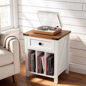 May include: A white wooden end table with a natural wood-toned top, featuring one drawer with a black knob and three open vertical storage compartments filled with vinyl records. A wood-grain record player with a clear plastic lid sits on top.